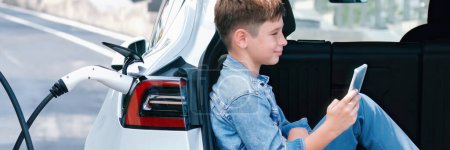 Photo for Little boy sitting on car trunk, using smartphone while recharging eco car from EV charging station. EV car road trip travel as alternative vehicle using sustainable energy concept.Panorama Perpetual - Royalty Free Image