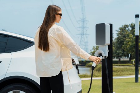 Jeune femme recharger la batterie de voiture EV à la station de charge connectée à l'installation industrielle électrique de tour de réseau électrique comme industrie électrique pour l'utilisation de véhicule respectueuse de l'environnement. Expedient