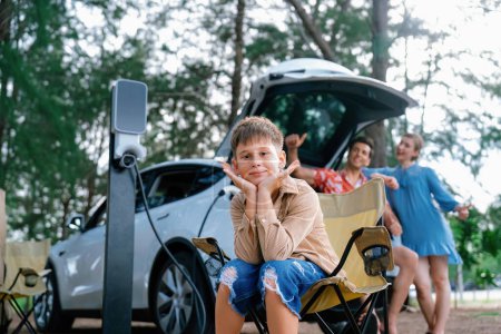 Photo for Little boy portrait sitting on camping chair with his family in background. Road trip travel with alternative energy charging station for eco-friendly car concept. Perpetual - Royalty Free Image
