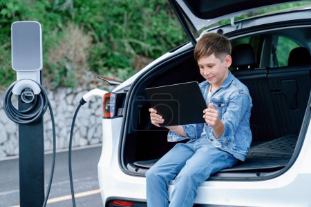 Photo for Little boy sitting on car trunk, using tablet while recharging eco-friendly car from EV charging station. EV car road trip travel as alternative vehicle using sustainable energy concept. Perpetual - Royalty Free Image