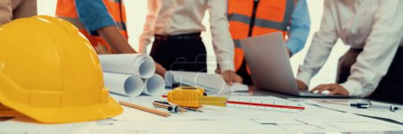 Photo for Engineer tool, architecture blueprint design and safety helmet hardhat on meeting table with contractors paper project with blurred engineer and architect working and discussing in background. Insight - Royalty Free Image