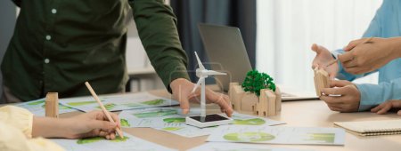 Photo for Windmill model represented renewable energy and wooden block represented eco city was placed on green business meeting table with environmental document scatter around. Front view. Delineation. - Royalty Free Image