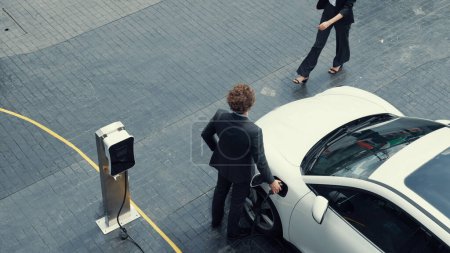 Photo for Progressive businessman and businesswoman leaning on electric car connected to charging station before driving around city center. Eco friendly rechargeable car powered by clean energy. - Royalty Free Image