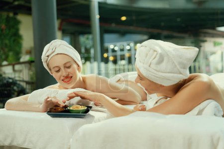 Photo for Couple of beautiful young girls lie on spa bed during interested in homemade beauty facial mask. Attractive woman touching herbal facial mask. Surrounded with nature environment. Tranquility. - Royalty Free Image