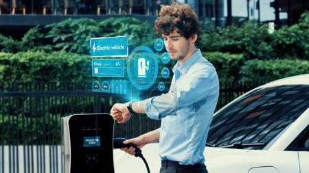 Foto de Hombre de negocios comprobar el estado de la batería del coche EV en el holograma smartwatch mientras se recarga desde el aparcamiento de la ciudad de la estación de carga. Estilo de vida futurista de energía limpia para la adaptación tecnológica de vehículos eléctricos. Período - Imagen libre de derechos