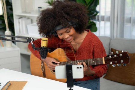 Foto de Anfitrión de canal en el músico de jóvenes afroamericanos tocando la guitarra junto con el canto, la radiodifusión en el teléfono inteligente en el estudio. Decoración de los auriculares del equipo y micrófono de grabación. Tastemaker. - Imagen libre de derechos