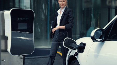 Photo for Progressive businesswoman unplug charger plug from charging station to his electric car before driving around city center. Eco friendly rechargeable car powered by sustainable and clean energy. - Royalty Free Image