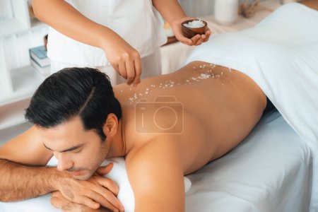 Photo for Blissful man customer having exfoliation treatment in luxury spa salon with warmth candle light ambient. Salt scrub beauty treatment in health spa body scrub. Quiescent - Royalty Free Image