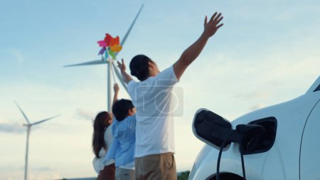 Téléchargez les photos : Concept de famille heureuse progressive appréciant leur temps au parc éolien avec véhicule électrique. Véhicule électrique alimenté par de l'énergie renouvelable propre provenant d'éoliennes pour la station de recharge. - en image libre de droit