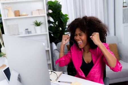 Téléchargez les photos : Blogueuse africaine visage extrêmement heureux, regardant à l'écran avec une réalisation appréciée à haut profit avec le plus récent projet de l'entreprise. Concept de travail d'expression gai à partir des modes de vie à la maison. Tastemaker. - en image libre de droit