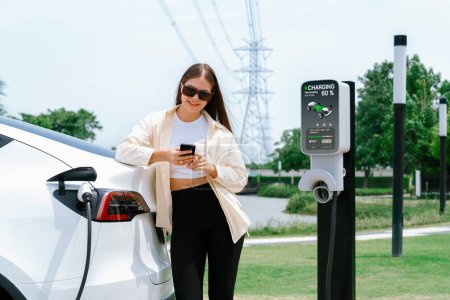 Foto de Mujer joven utiliza el teléfono inteligente para pagar la electricidad en la estación de carga de coches EV público parque verde de la ciudad. Moderno estilo de vida urbano ambiental y sostenible con vehículo EV. Expediente - Imagen libre de derechos