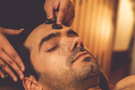 Téléchargez les photos : Homme caucasien appréciant relaxant massage de la tête anti-stress avec pierre chaude et dorloter beauté faciale loisirs de la peau dans la lumière chaude bougie salon de spa ambiant dans un complexe ou un hôtel. Tranquillité - en image libre de droit