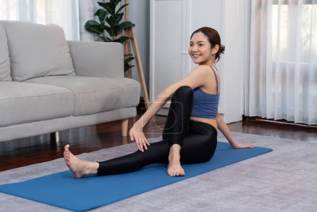 Foto de Mujer asiática en ropa deportiva haciendo ejercicio de yoga en la alfombra de fitness como su rutina de entrenamiento en casa. Cuidado corporal saludable y meditación tranquila en el estilo de vida del yoga con comodidad y relajación. Vigoroso - Imagen libre de derechos