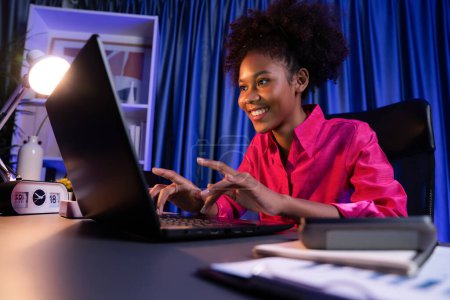 Téléchargez les photos : Blogueuse africaine portant une chemise rose avec un visage heureux, regardant un ordinateur portable à l'écran avec un projet de réussite valorisé ou obtenir une bourse d'études. Concept de travail d'expression gai de la maison. Tastemaker. - en image libre de droit