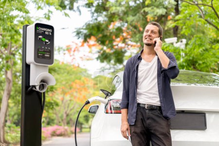 Photo for Man talking on smartphone while recharging electric car battery charging from EV charging station during vacation holiday road trip at national park or autumnal forest. Exalt - Royalty Free Image