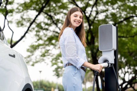Photo for Young woman recharge EV electric vehicles battery from EV charging station in outdoor green city park scenic. Eco friendly urban transport and commute with eco friendly EV car travel. Exalt - Royalty Free Image