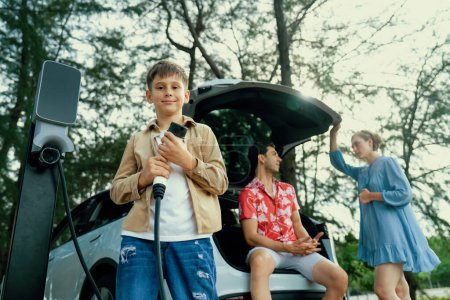 Photo for Little boy holding EV charger and point at camera with his family sitting on the trunk in background. Road trip travel with alternative energy charging station for eco-friendly car concept. Perpetual - Royalty Free Image