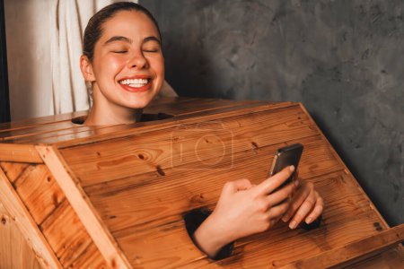 Téléchargez les photos : Un portrait de la magnifique femme caucasienne jouant son téléphone portable tout en utilisant une armoire de sauna en bois dans un ton chaud. Jolie femelle avec une belle peau prenant une photo. Fond gris. Tranquillité - en image libre de droit