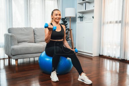 Photo for Athletic and sporty senior woman sitting on fit ball while engaging in weight lifting with dumbbell at home exercise as concept of healthy fit body lifestyle after retirement. Clout - Royalty Free Image