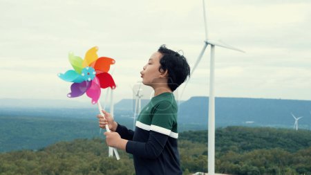 Foto de Progresivo joven asiático jugando con el juguete de la turbina de viento en el parque eólico, campo verde sobre la colina. Energía verde procedente del generador eólico eléctrico renovable. - Imagen libre de derechos