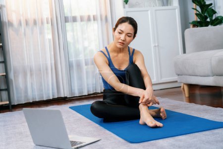 Téléchargez les photos : Femme asiatique en vêtements de sport assis sur le sol de la maison tout en regardant la vidéo de formation d'exercice en ligne sur ordinateur portable. Fille attrayante s'engager dans sa poursuite d'un mode de vie sain. Vigoureux - en image libre de droit