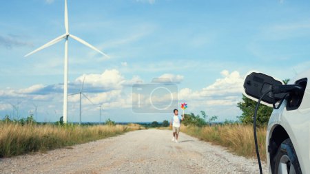 Photo for Progressive young boy playing windmill toy next to EV car at wind turbine farm. Electric generator from wind by wind turbine generator on the country side. - Royalty Free Image