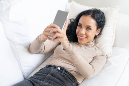 Photo for Relaxed young African American woman using crucial mobile phone on sofa couch in living room at home - Royalty Free Image