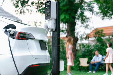 Photo for Focus electric vehicle recharge from home charging station on blur background of happy and playful family playing together. EV car using alternative and sustainable energy for better future.Synchronos - Royalty Free Image