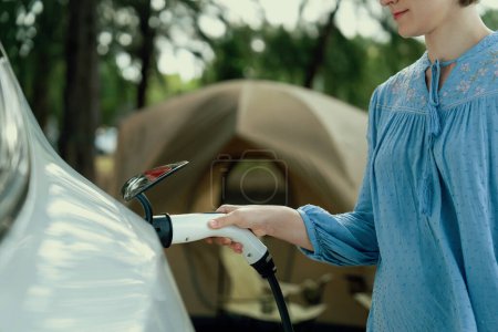 Photo for Holiday camping vacation traveling to nature countryside with electric car, woman recharge EV car from charging station on campsite. Natural travel and eco car for sustainable environment. Perpetual - Royalty Free Image