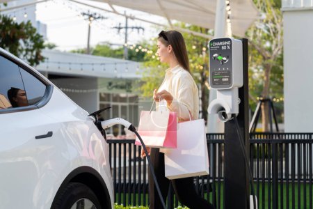 Photo for Young woman holding shopping bag recharge EV car battery from charging station at parking lot. Modern woman go shopping by environmental friendly electric vehicle in urban travel lifestyle. Expedient - Royalty Free Image