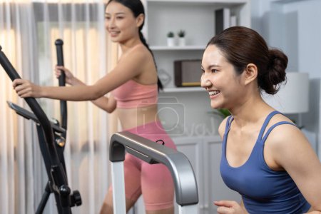 Foto de Mujer asiática atlética enérgica y fuerte corriendo en máquina elíptica en casa con compañero de entrenamiento o entrenador. Búsqueda de físico en forma y compromiso con un estilo de vida saludable. Vigoroso - Imagen libre de derechos