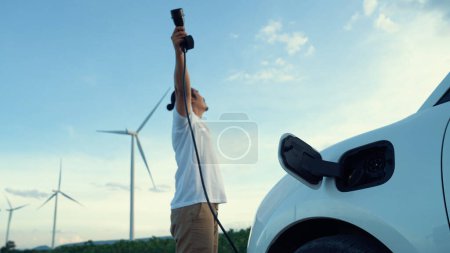 Foto de Hombre progresivo con su coche eléctrico, coche EV recarga energía de la estación de carga en el campo verde con turbina eólica como concepto de energía sostenible futuro. Vehículo eléctrico con generador de energía. - Imagen libre de derechos