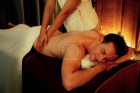 Foto de Hombre caucásico cliente disfrutando de relajante masaje de spa anti-estrés y mimos con belleza recreación de la piel de ocio en caliente iluminación de velas salón de ambiente spa en complejo de lujo u hotel. Silencioso. - Imagen libre de derechos