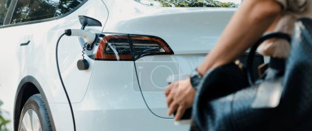 Photo for Young man recharge electric cars battery from charging station in outdoor green city park. Rechargeable EV car for sustainable environmental friendly urban travel. Panorama Expedient - Royalty Free Image