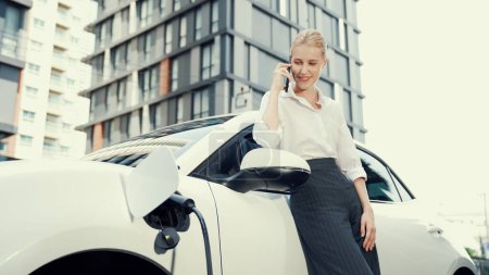 Photo for Progressive businesswoman leaning on electric car and charging station before driving around city center. Eco friendly rechargeable EV car powered by sustainable and clean energy. - Royalty Free Image