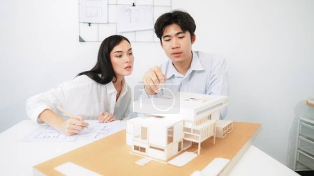 Photo for Closeup of skilled architecture engineer team measure and decide house model while working together and discussing about house structure at meeting room with blueprint placed behind. Immaculate. - Royalty Free Image