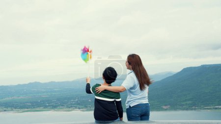 Foto de Una mujer progresista y su hijo están de vacaciones, disfrutando de la belleza natural de un lago en el fondo de una colina mientras el niño lleva un molino de viento de juguete. - Imagen libre de derechos