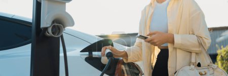 Photo for Woman holding shopping bag and use smartphone to pay for electricity to recharge EV car battery from charging station at city mall parking lot. Modern woman go shopping by eco car.Panorama Expedient - Royalty Free Image