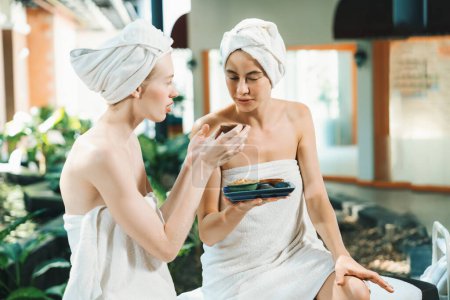 Photo for Couple of beautiful woman interested in homemade facial masks while sitting at spa salon. Attractive woman in white towel enjoy herbal masks with her friend surrounded by nature. Tranquility. - Royalty Free Image