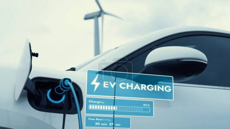 Photo for Electric car recharging energy from EV charging station display futuristic smart battery status hologram by EV charger plug cable in wind turbine farm. Alternative clean energy sustainability. Peruse - Royalty Free Image