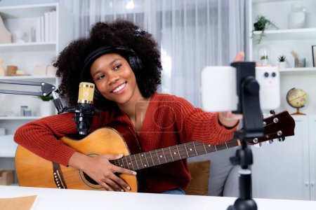 Photo for Host channel of beautiful African woman influencer setting smartphone, sing with play guitar in broadcast. Time slot of music blogger on live social media online. Concept of audio creator. Tastemaker. - Royalty Free Image