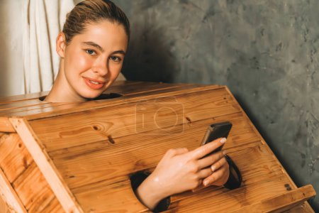 Photo for Pretty caucasian girl looking at camera while playing her mobile phone at sauna room. Attractive female with beautiful skin looking at camera during using wooden sauna cabinet. Tranquility. - Royalty Free Image