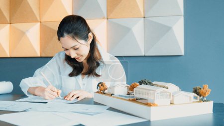 Photo for Portrait of beautiful young architect engineer measures house model while taking a note on blueprint with laptop, project plan and blueprint placed on table. Business design concept. Manipulator. - Royalty Free Image