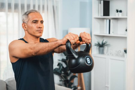 Photo for Athletic and sporty senior man engaging in body workout routine with lifting kettlebell at home as concept of healthy fit body with body weight lifestyle after retirement. Clout - Royalty Free Image