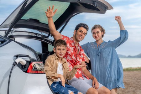 Photo for Family vacation trip traveling by the beach with electric car, lovely family sit on the trunk, charging EV car battery with green and sustainable energy. Family travel and eco-friendly car. Perpetual - Royalty Free Image