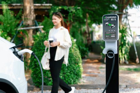 Jeune femme et trajets urbains durables avec la recharge de voiture électrique EV au café extérieur dans le jardin printanier, la durabilité de la ville verte et la voiture écologique EV. Expedient