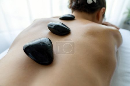 Téléchargez les photos : Massage aux pierres chaudes au salon de spa dans un complexe de luxe avec lumière du jour sérénité ambiante, femme heureuse client profitant d'un massage aux pierres de basalte spa glisser sur le corps avec une chaleur apaisante. Tranquillité - en image libre de droit