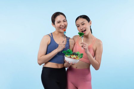 Photo for Two young sporty Asian women in sportswear holding salad bowl fill with fruit and vegetable. Natural youthful and fit body lifestyle people with balance nutrition on isolated background. Vigorous - Royalty Free Image