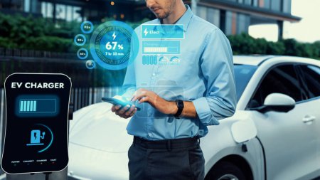 Photo for Businessman checking battery status hologram from EV smartphone application while his EV car recharging from charging station in residential area. Futuristic clean sustainable EV tech. Peruse - Royalty Free Image