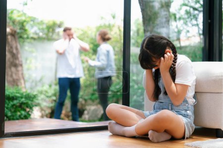 Photo for Stressed and unhappy young girl huddle in corner, cover her ears blocking sound of her parent arguing in background. Domestic violence at home and traumatic childhood develop to depression. Synchronos - Royalty Free Image